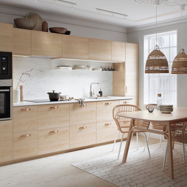 Een landelijke keuken met houten frontjes en ee RINDSHOLM vloerkleed op de grond. Met een houten eettafel, rotan eetkamerstoelen en twee rotan hanglampen aan het plafond.