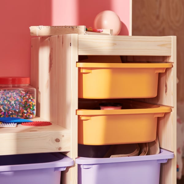 In einem weiß gebeizten TROFAST Regalrahmen in Kiefer befinden sich hellorangefarbene und lilafarbene Boxen voller Kunstmaterialien und Spielzeug.