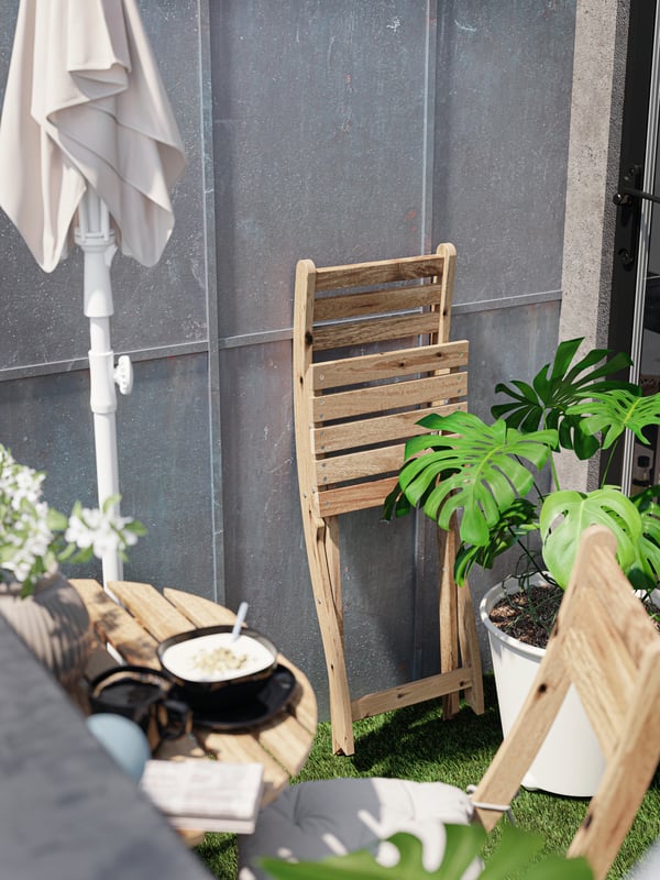 Una silla de madera plegada contra una pared de metal y una sombrilla de color beige grisáceo plegada, una maceta blanca y un desayuno sobre una mesa pequeña.