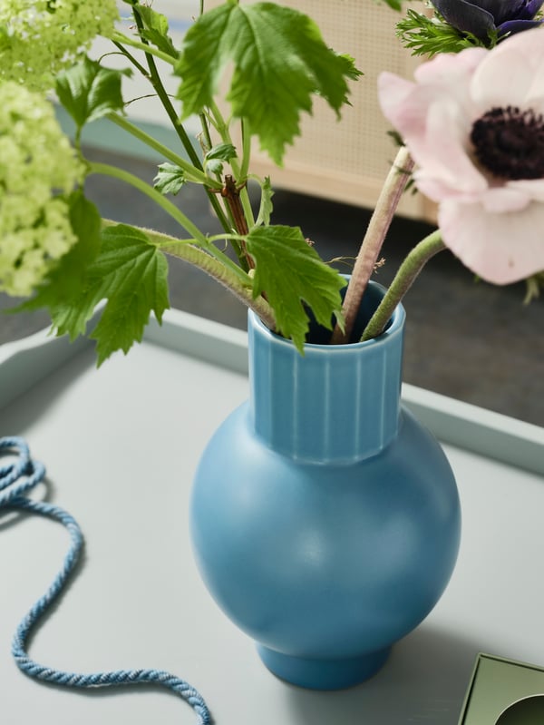 A blue OMFÅNG vase with a small bouquet made of fresh and artificial flowers and greenery standing on a tray.