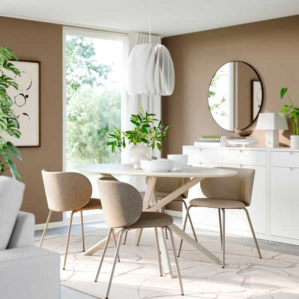 Cette salle à manger IKEA lumineuse comprend une table ronde blanche, quatre chaises beiges, une suspension sculpturale, un buffet blanc avec un miroir rond et de luxuriantes plantes vertes baignés de lumière naturelle.