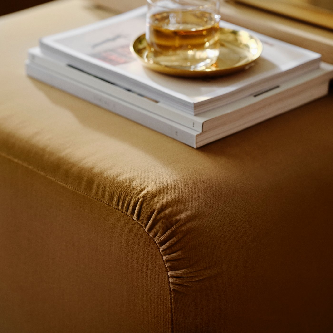 Una ciotola e un piatto STOCKHOLM 2025 color ottone posti su alcuni libri e su un pouf Alhamn beige.