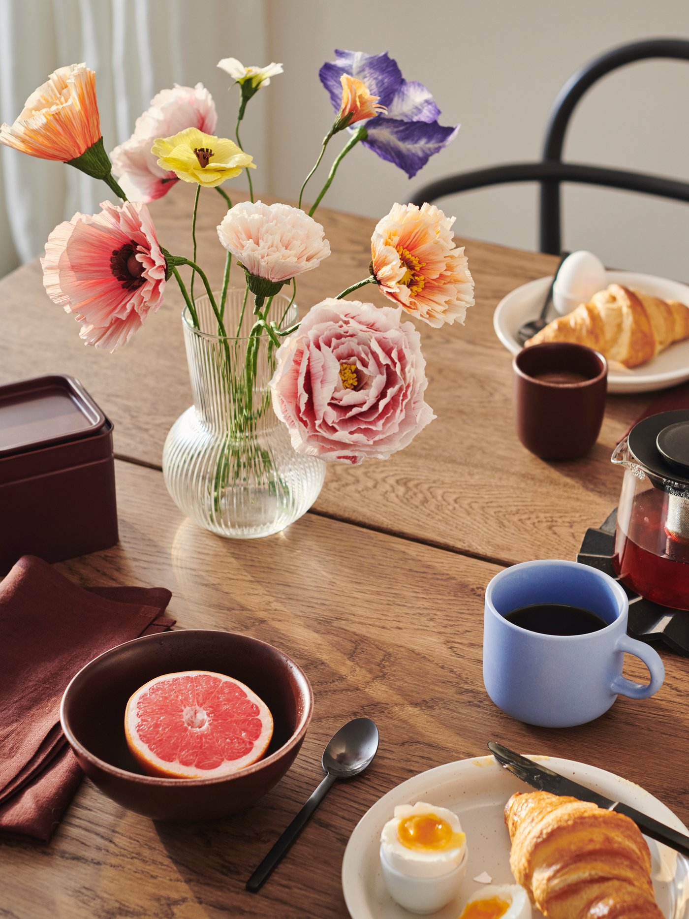 A PÅDRAG vase containing a number of GULDSPETT artificial flowers stands on a table that is set for breakfast.