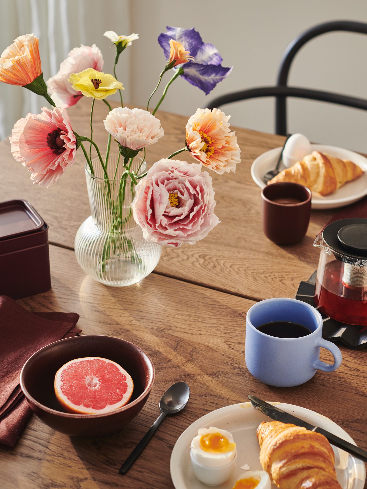 A PÅDRAG vase containing a number of GULDSPETT artificial flowers stands on a table that is set for breakfast.