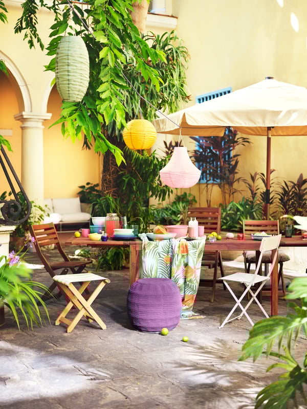 A wooden table and mis-matched chairs stand outside against a yellow wall, with an umbrella and colourful lanterns.