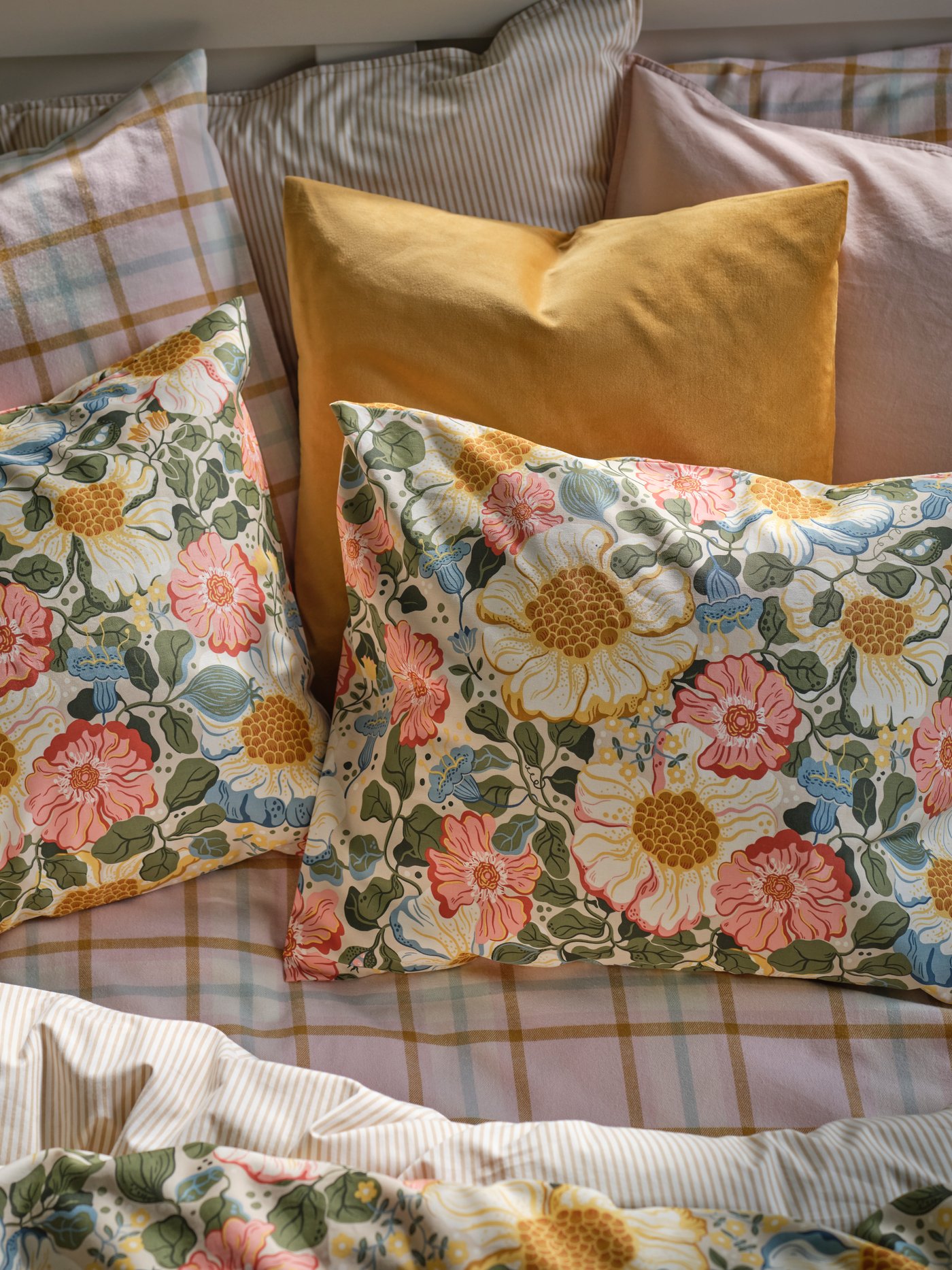 A bed with pillows and duvets in ARENBOK bed linen in beige and floral combined with yellow, pink and chequered pillows.