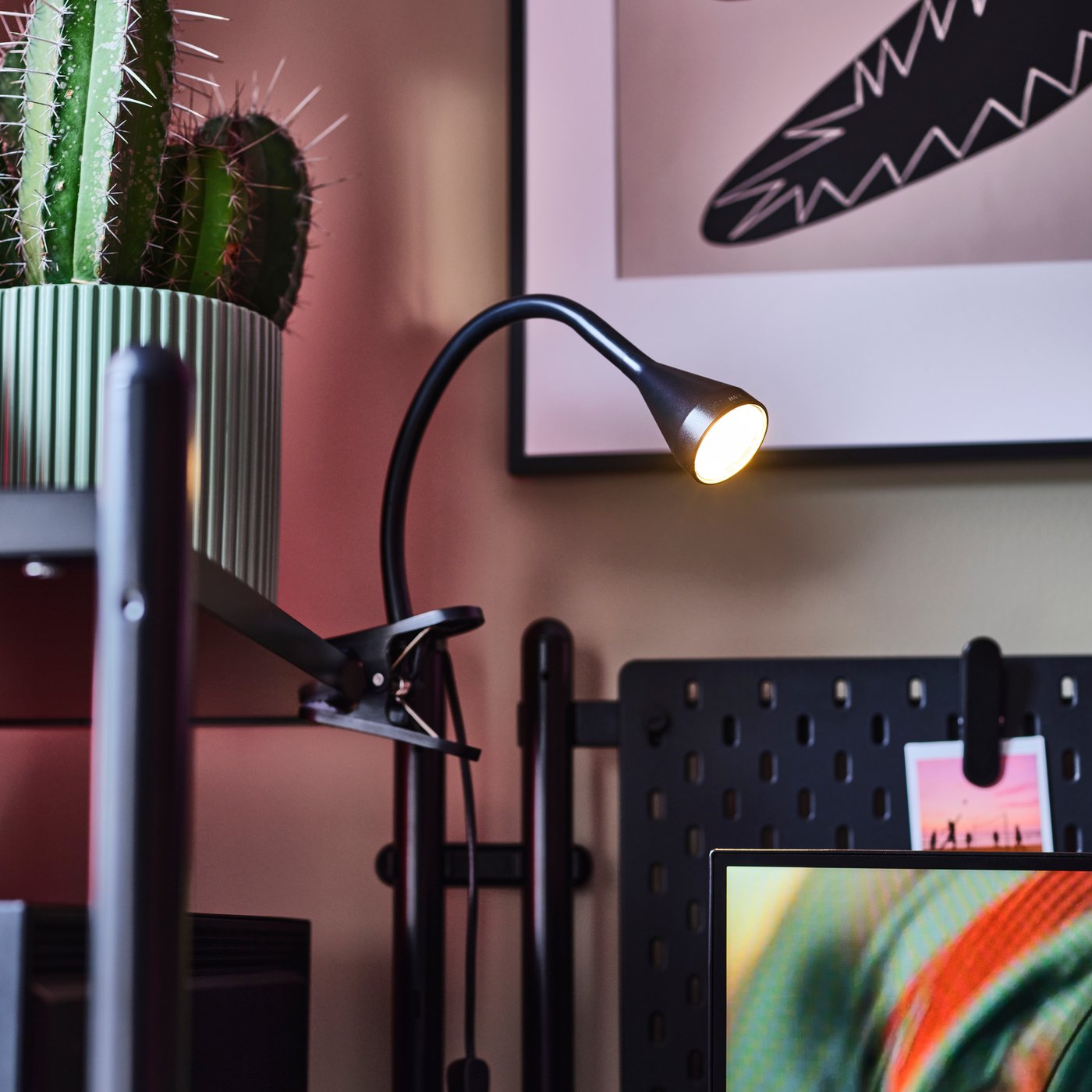 A workspace with a black NÄVLINGE LED clamp spotlight shining on a desk alongside a vibrant green cactus in a pot.