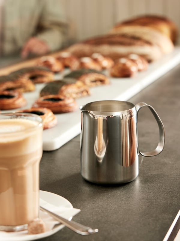 A close-up of a silver MATTLIG milk jug sitting on a table beside a clear POKAL glass filled with coffee.