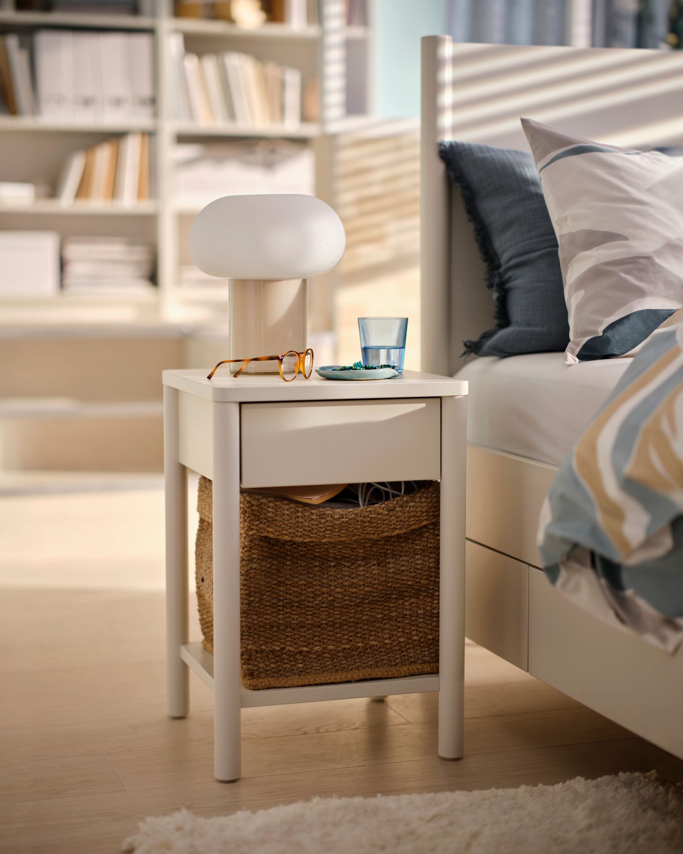 White bedside table next to a white bed, with a basket on the lower shelf, a table lamp and glass of water on top.
