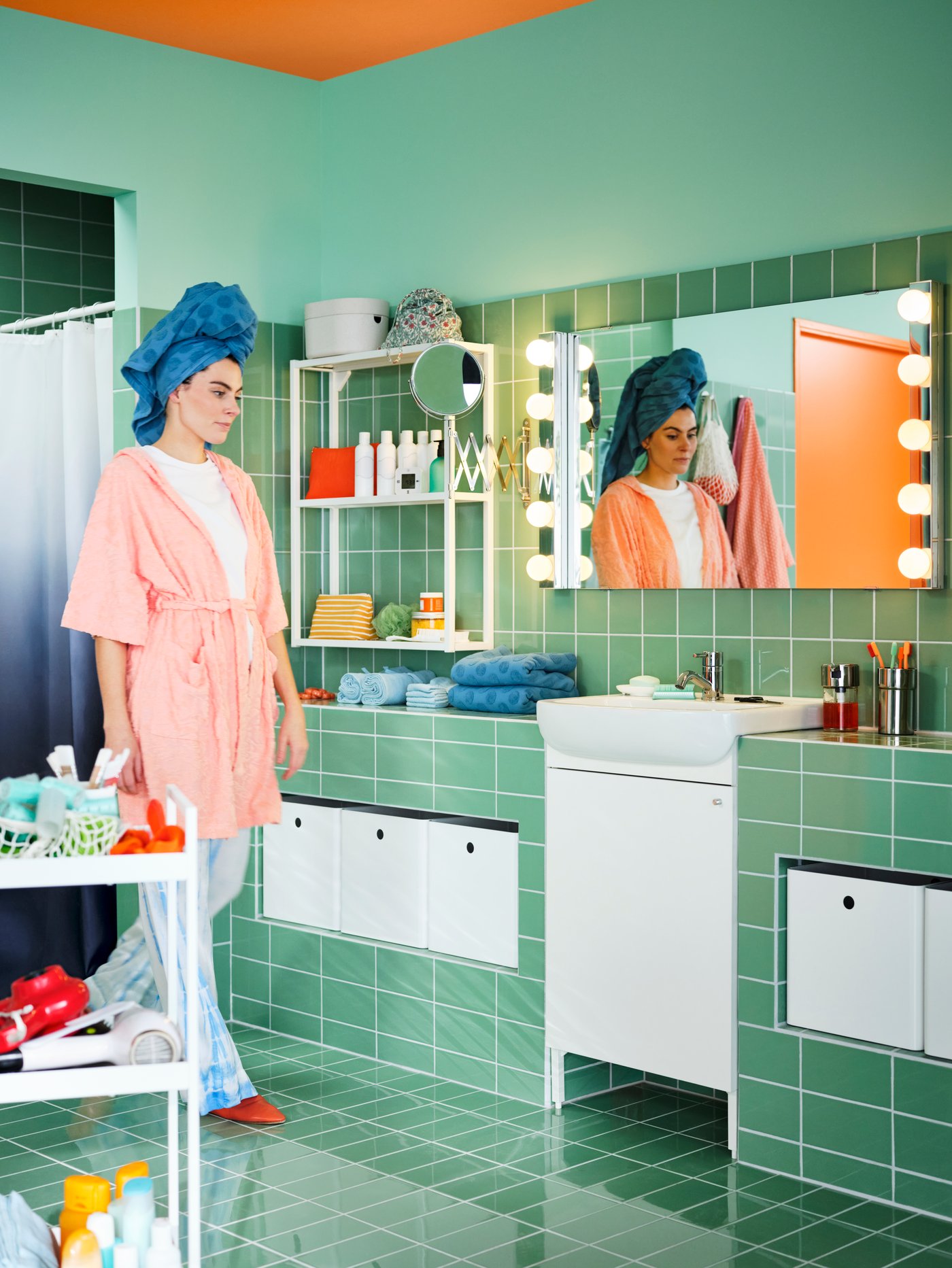 A green-tiled bathroom with a white BJÖRKÅN sink and NYSJÖN wash basin cabinet with one door and a woman wearing pyjamas.
