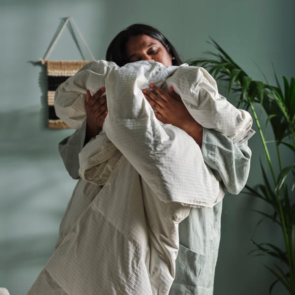 A bedroom with a person holding a warm FJÄLLHAVRE duvet, surrounded by wall decor and a green plant.