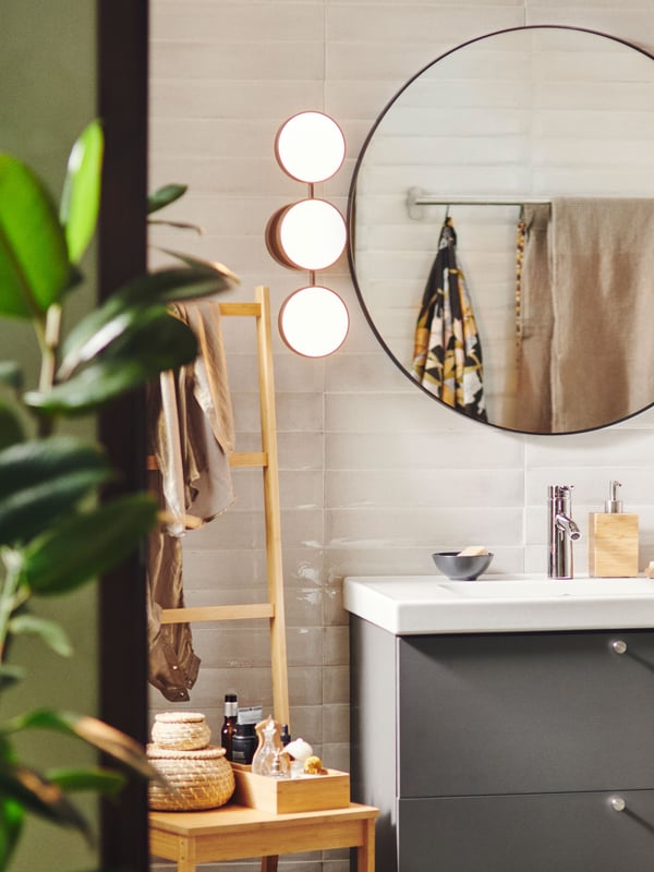 A dimmable matte black KABOMBA LED wall lamp is placed next to a round FAXÄLVEN mirror above a grey sink vanity in a bathroom.