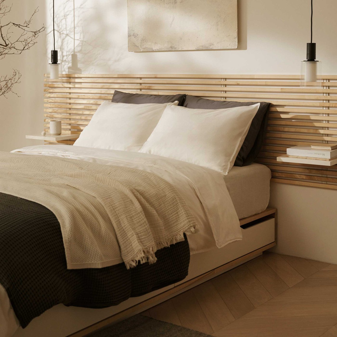 穏やかで居心地の良いベッドルームは、安らかな眠りにぴったりです。快適なベッドには、明るい木製のスラットヘッドボードと一体型の棚が備わり、夜の必需品を置くのに便利です。清潔感のある白い寝具、柔らかなダークグレーの枕、温かみのあるニュートラルトーンの心地よい質感のスローが、美しく調和しています。両サイドには洗練されたペンダントライトが優雅に吊り下げられ、柔らかな間接照明を演出しています。ベッドの上には、ミニマルな抽象画が落ち着いた雰囲気を添えています。温かみのある木製の部屋と小さなダークカラーのラグが、調和のとれたリラックスした雰囲気を演出しています。