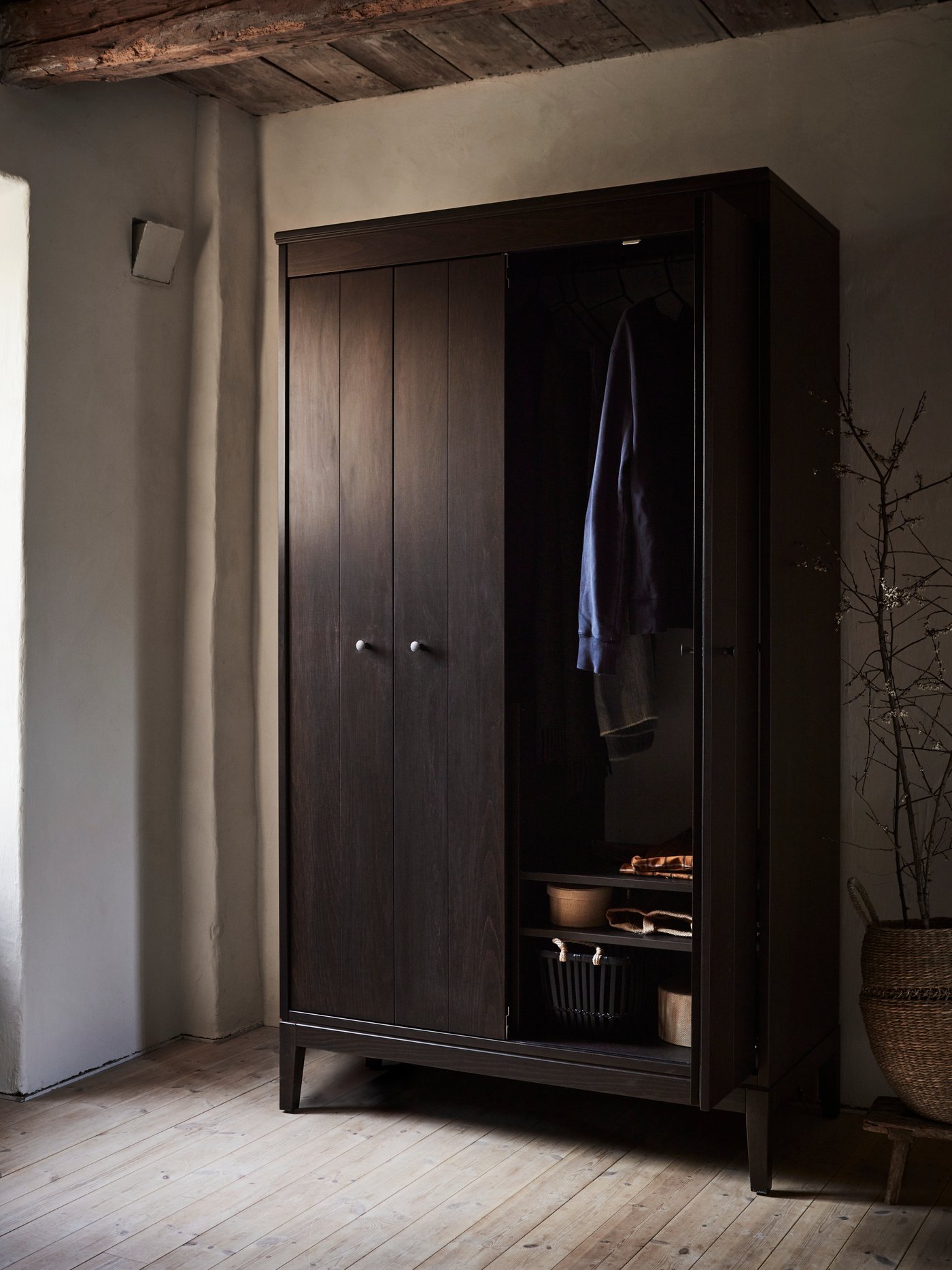 A dark brown stained IDANÄS wardrobe in a rustic style bedroom with the right hand door open and clothes on the hanging rail.