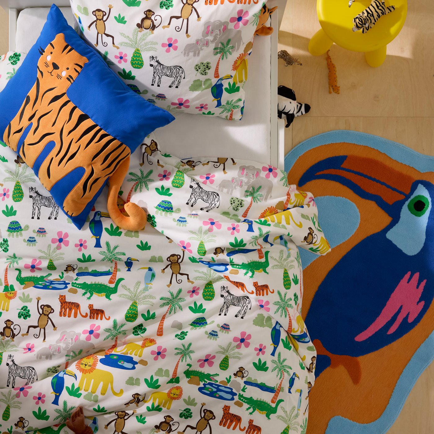 A vista d'ocell, llit infantil blanca amb roba de llit de colors de temàtica selvàtica, coixí amb motiu de tigre i catifa de tucà.