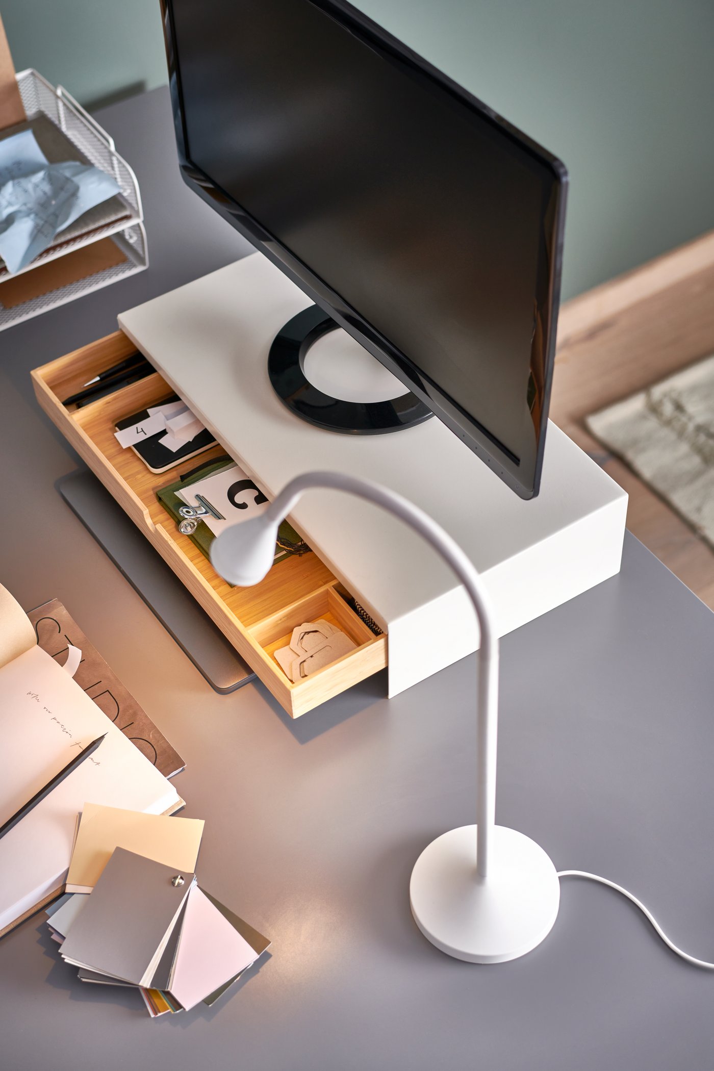 A white NÄVLINGE LED work lamp on a grey table top, beside an ELLOVEN monitor stand with a drawer that’s open.