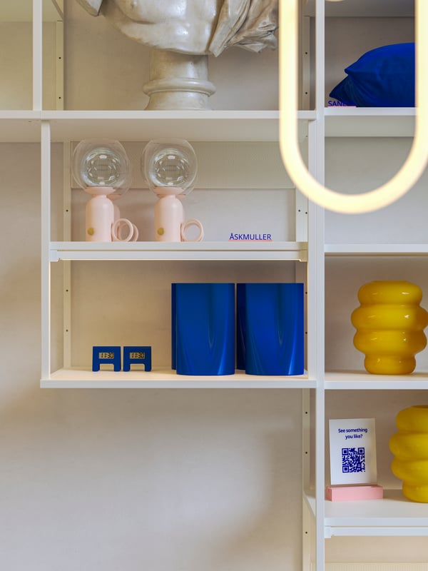 Wall-mounted white PLATSA open shelving units display bright yellow, blue and pale pink home deco objects and a QR-code sign. 