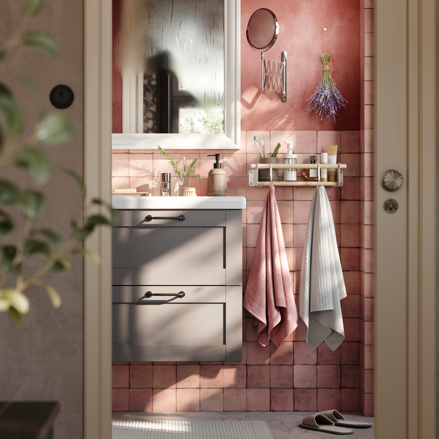 Casa de banho rosa com armário para lavatório com duas gavetas ENHET em cinzento, espelho em branco e duas toalhas penduradas numa calha com ganchos.