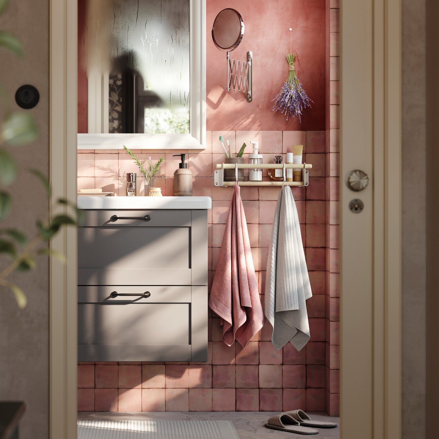 Es ist ein rosafarbenes Badezimmer mit einem grauen ENHET Waschbeckenschrank mit zwei Schubladen, einem weissen Spiegel und zwei Handtüchern zu sehen, die an einer Stange mit Haken hängen.