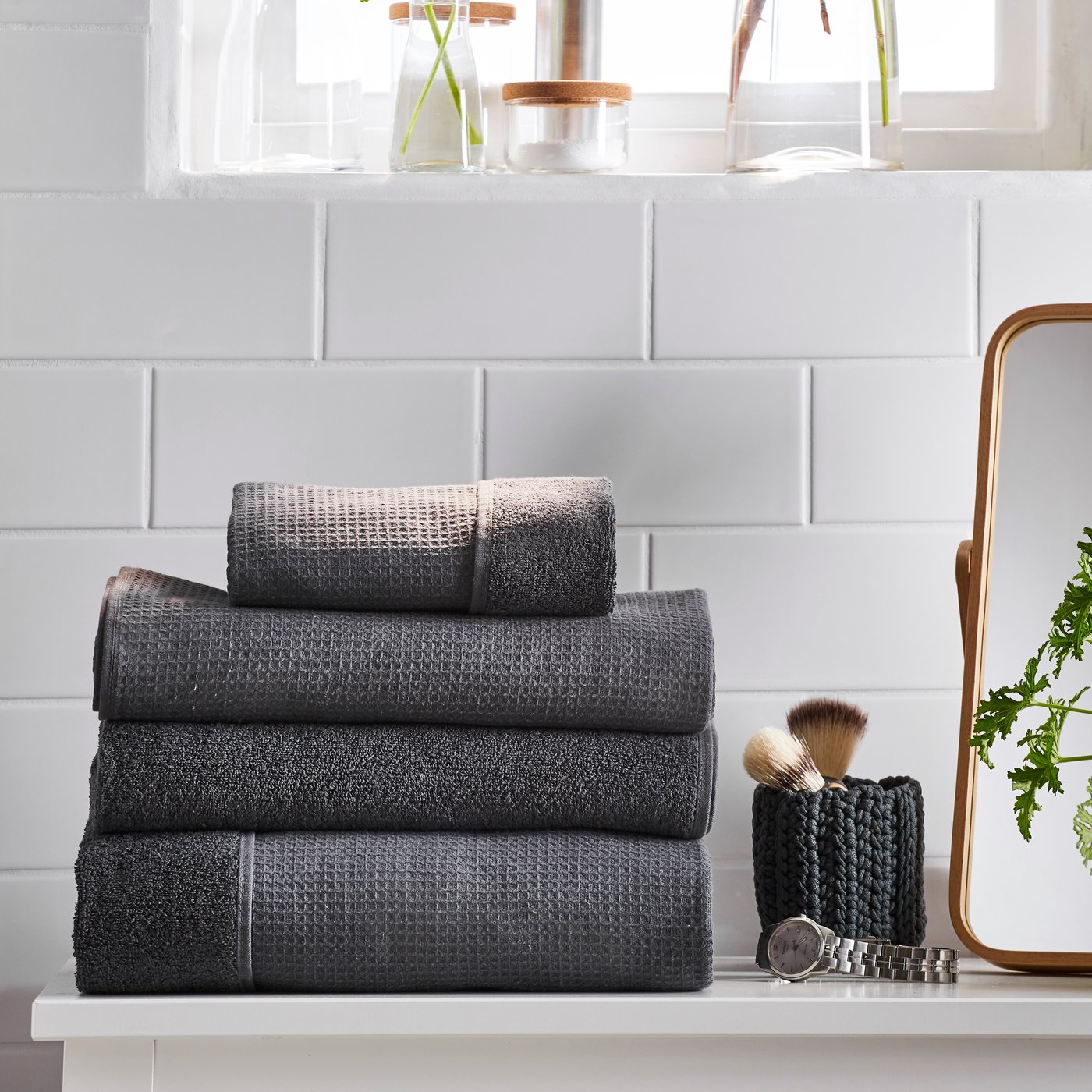 SALVIKEN hand towel and bath towel in anthracite next to watch and mirror in front of white tiles.