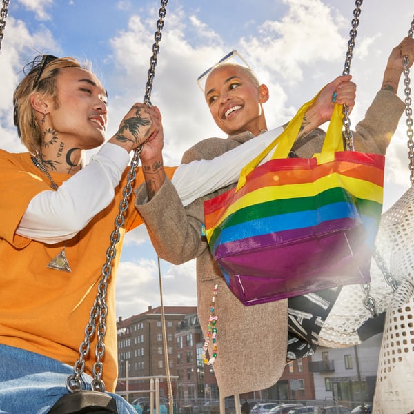 Two people outside riding on swings next to each other, one of them holds a small STORSTOMMA bag in their hand.