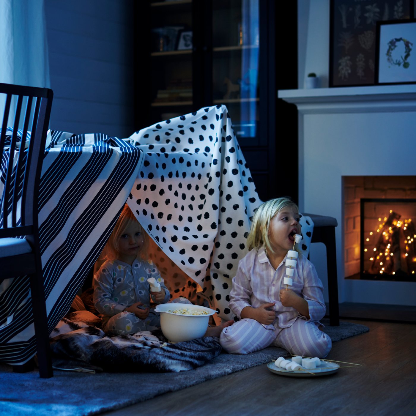Two young children are eating marshmallows while sitting on the floor in the living room inside a homemade hut or tent made from a tablecloth.
