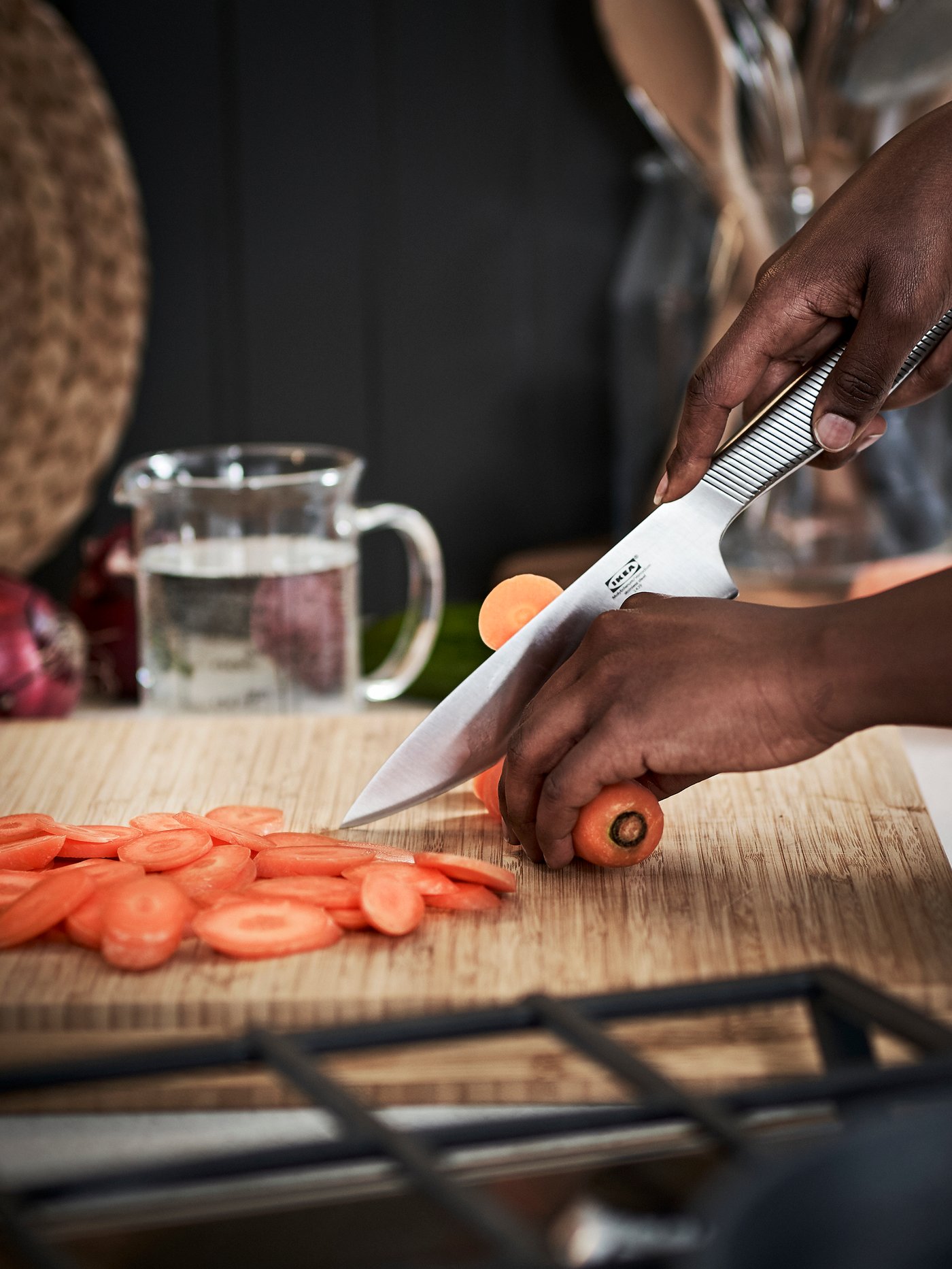Close up on kitchen counter with person cutting carrots with stainless steel utility knife on large wooden chopping board.