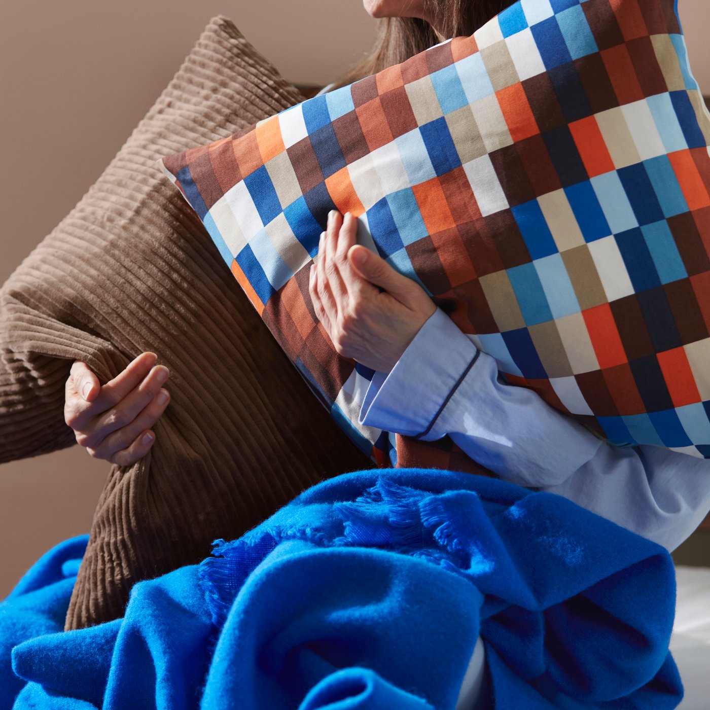 Una persona sostiene in braccio due grandi cuscini con fodera marrone e uno con tinta a rettangoli colorati