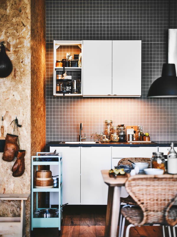 An ENHET kitchen sits next to a NISSAFORS trolley against the wall, with cooking gear. A dining table and chairs sit nearby.