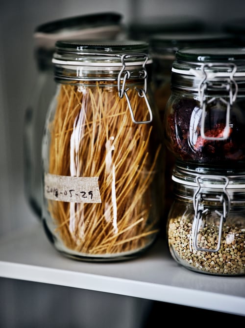 One large and two small glass KORKEN jars filled with dried fruit, pulses and pasta on a white shelf.