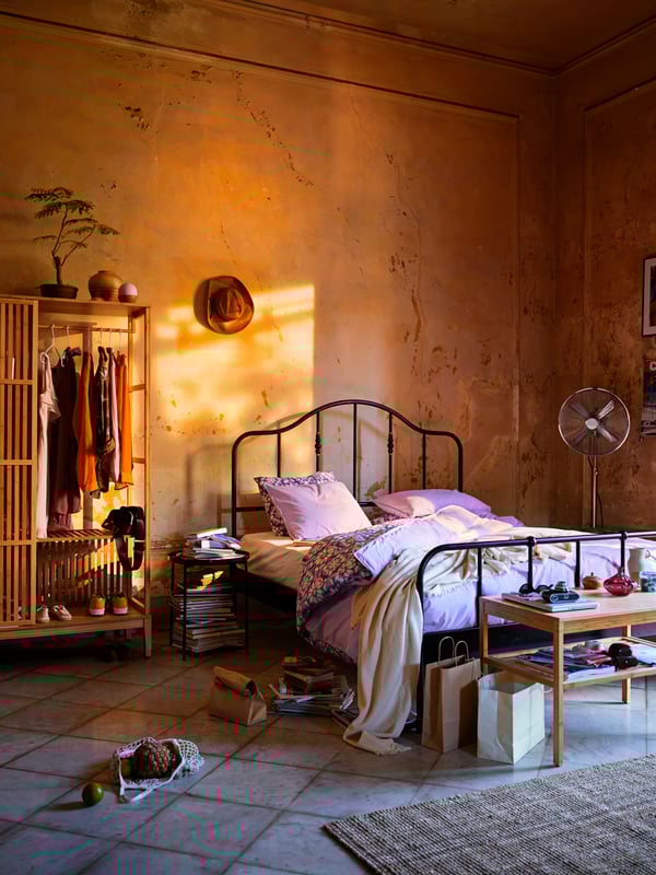 Schlafzimmer mit einem schwarzen Metallbett. Am Bettende steht eine schlichte Bettbank aus Holz. Neben dem Bett ist ein halb offener Kleiderschrank.