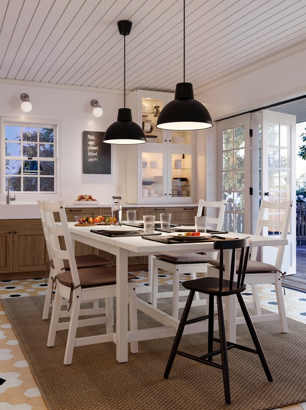 Une cuisine où se trouve une table NORDVIKEN blanche entourée de cinq chaises. Le repas est prêt à être servi. Au-dessus de la table sont fixées deux suspensions SKURUP