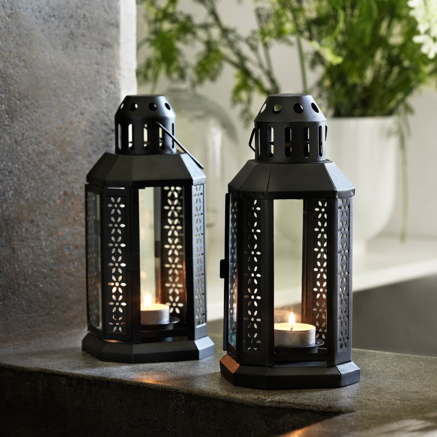 Two ENRUM lanterns for tea light, each containing a lit tea light, standing on a dark grey stone shelf.