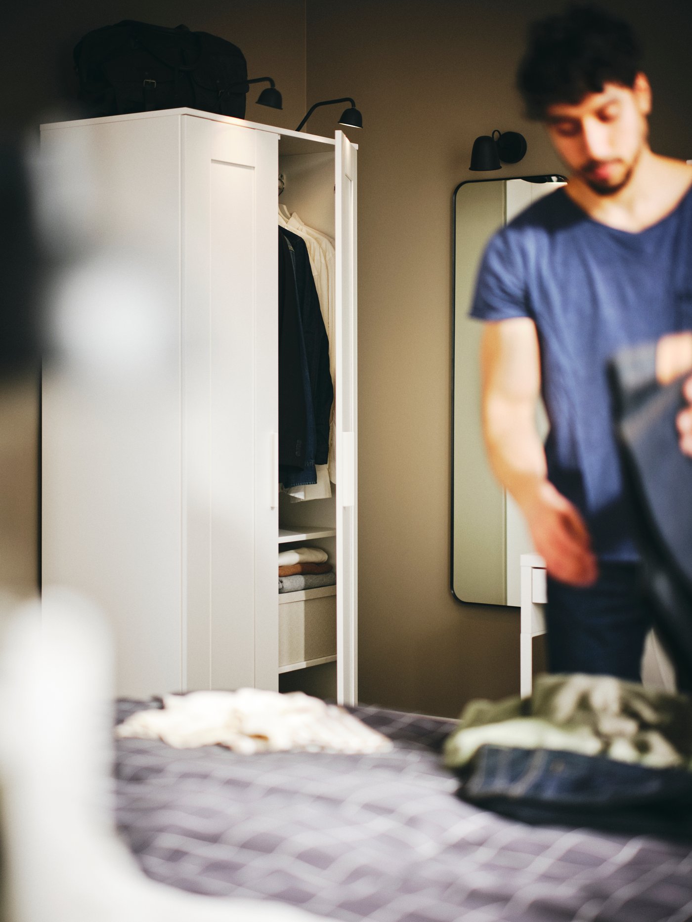 Dormitorio con una persona doblando la ropa junto a un clóset blanco con 2 puertas de madera, iluminado por 2 lámparas de clóset.