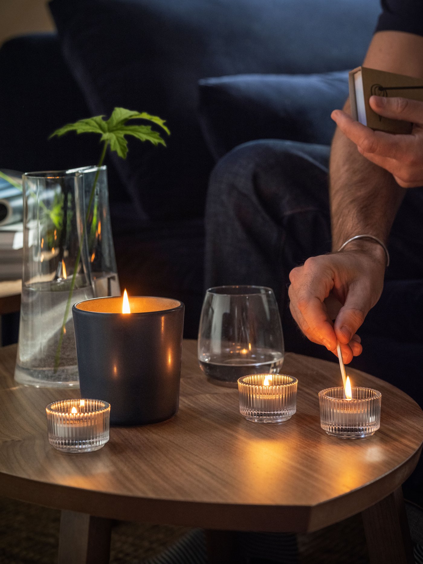 A vetiver & geranium/black-turquoise FRUKTSKOG scented candle in ceramic jar sits on a table next to other lit candles in clear candle holders.