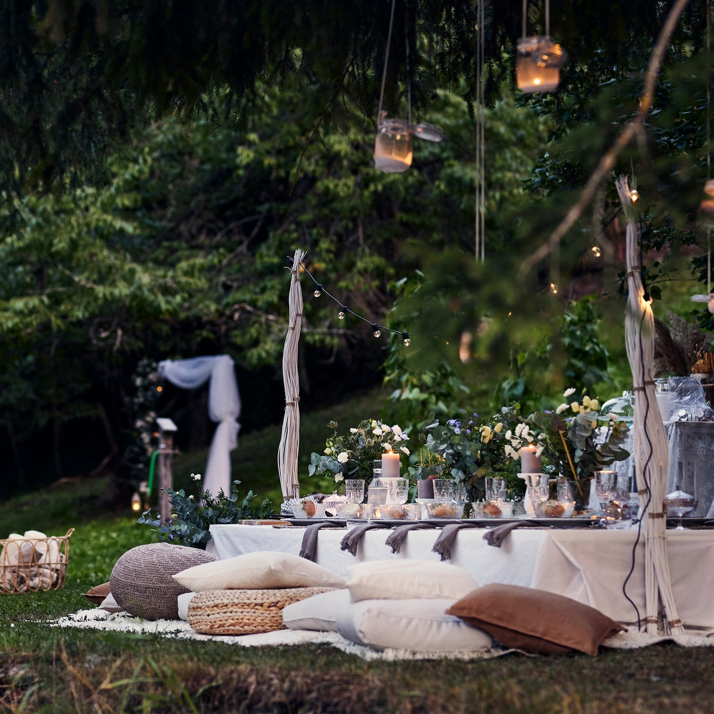 Ett långbord dukat för fest med puffar och kuddar som sittplatser i en trädgård under en sommarkväll.