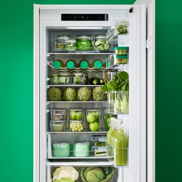An integrated SKOGNÄS fridge/freezer has different kinds of green vegetables and fruit and green containers stored inside.