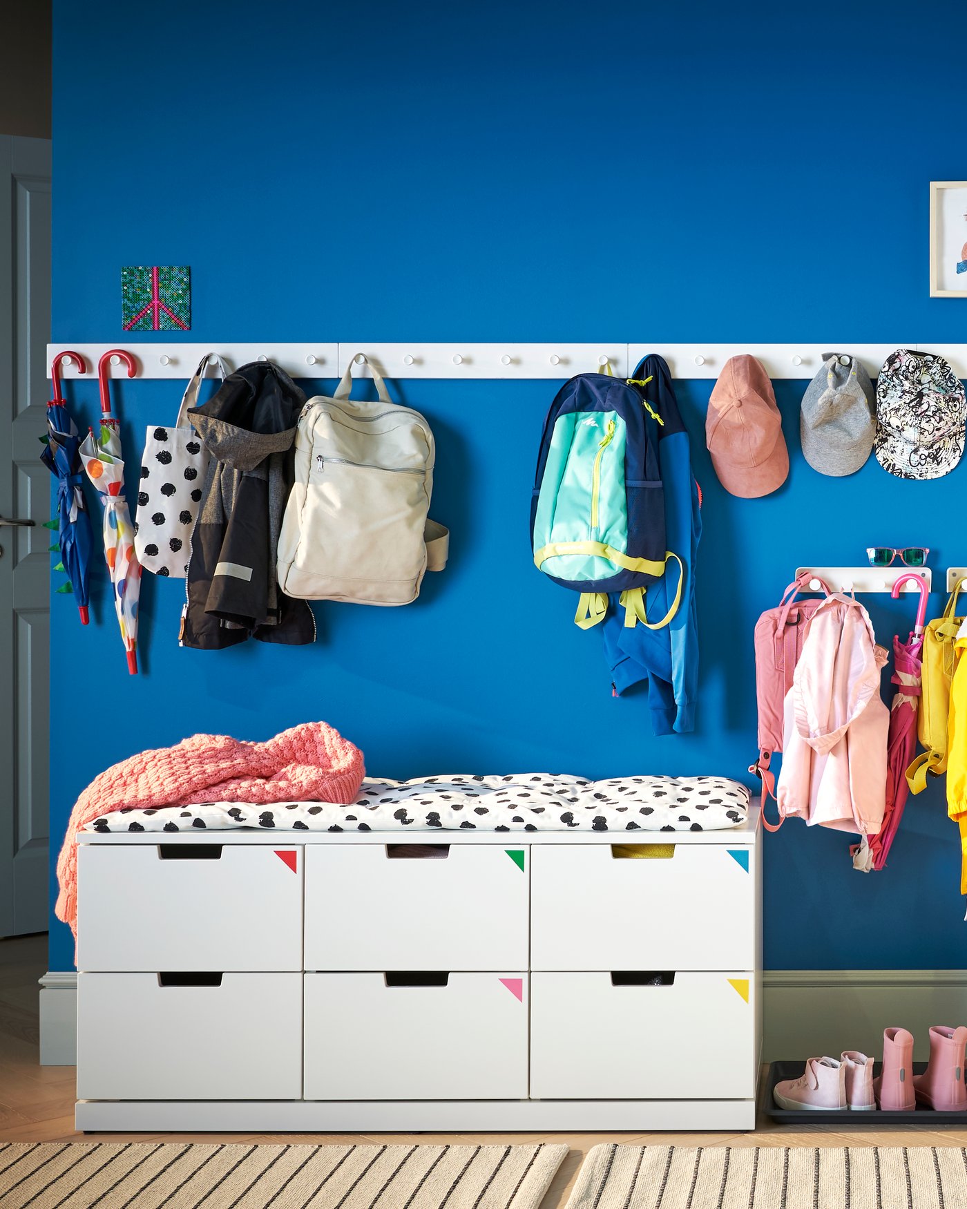 En vit låg NORDLI byrå med sittplats står framför en blå vägg med en vit dyna med svarta prickar på. Ovanför händer klädhängare med ryggsäckar och kepsar i olika färger.
