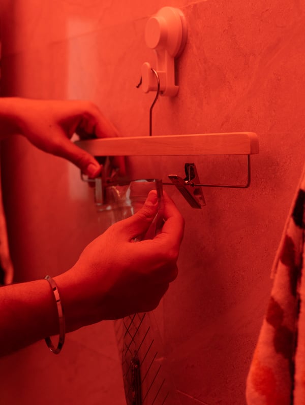 Two hands hang a BUMERANG skirt hanger on a TISKEN hook on a bathroom wall. The lighting is red.