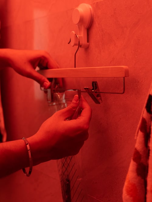 Zwei Hände befestigen einen ISTAD wiederverschließbaren Beutel an einem BUMERANG Rockbügel, der an einem TISKEN Haken mit Saugnapf hängt. In dem Beutel befindet sich ein Mobiltelefon.