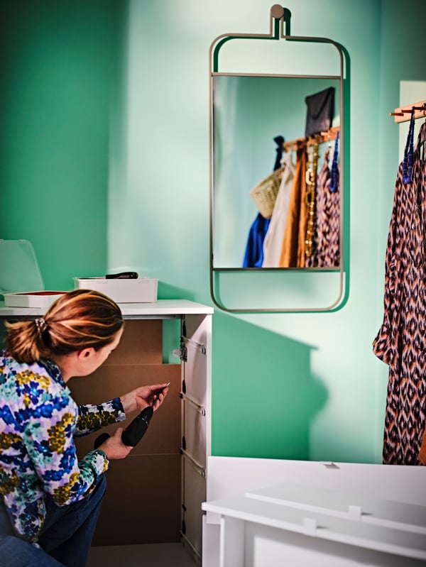 A person wall-anchoring a white VIHALS chest of 4 drawers, beside a green wall-hanging mirror and an oak rack with 6 knobs.