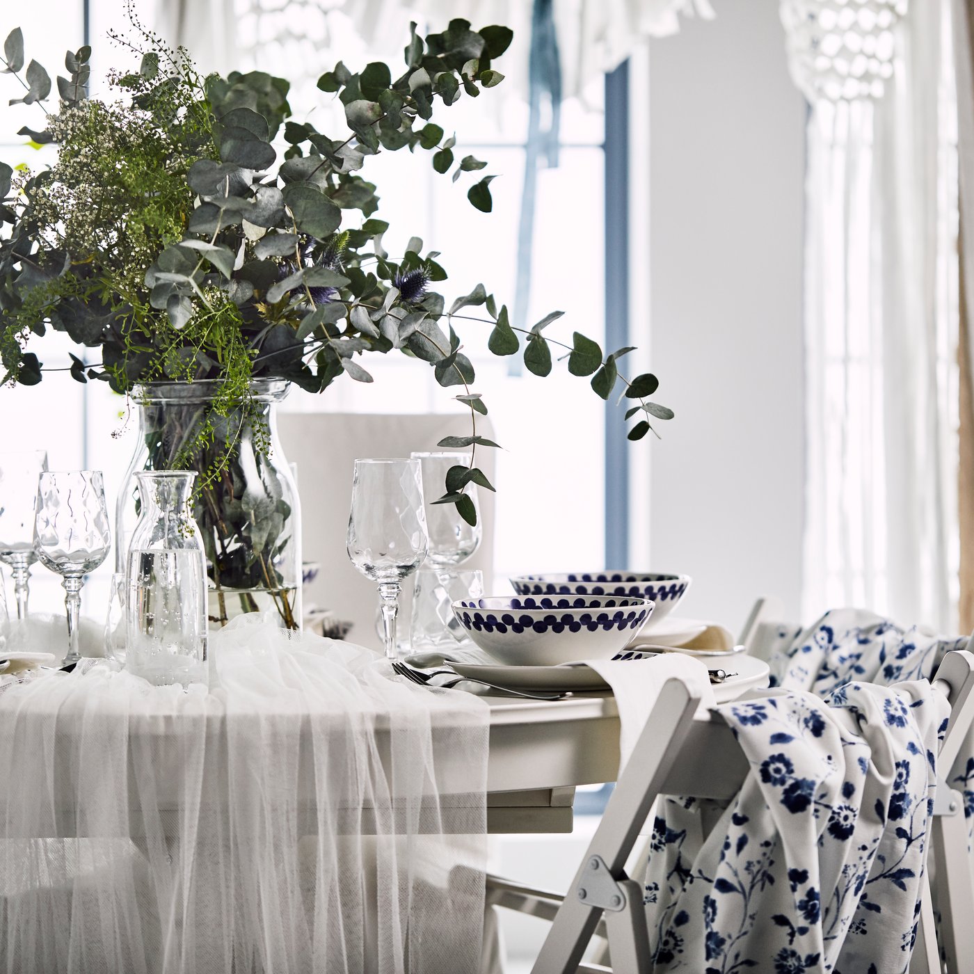 Mise en place romantica, con tulle bianco su un tavolo rotondo e un vaso con fiori di campo e foglie di eucalipto.