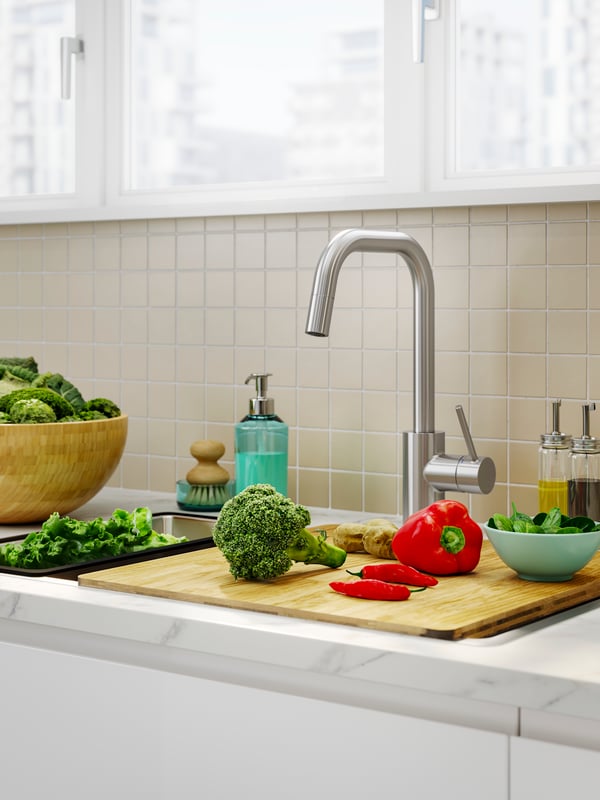 The NORRSJÖN chopping board holds broccoli, peppers, and chillies on a kitchen counter, next to a tap and kitchen essentials.