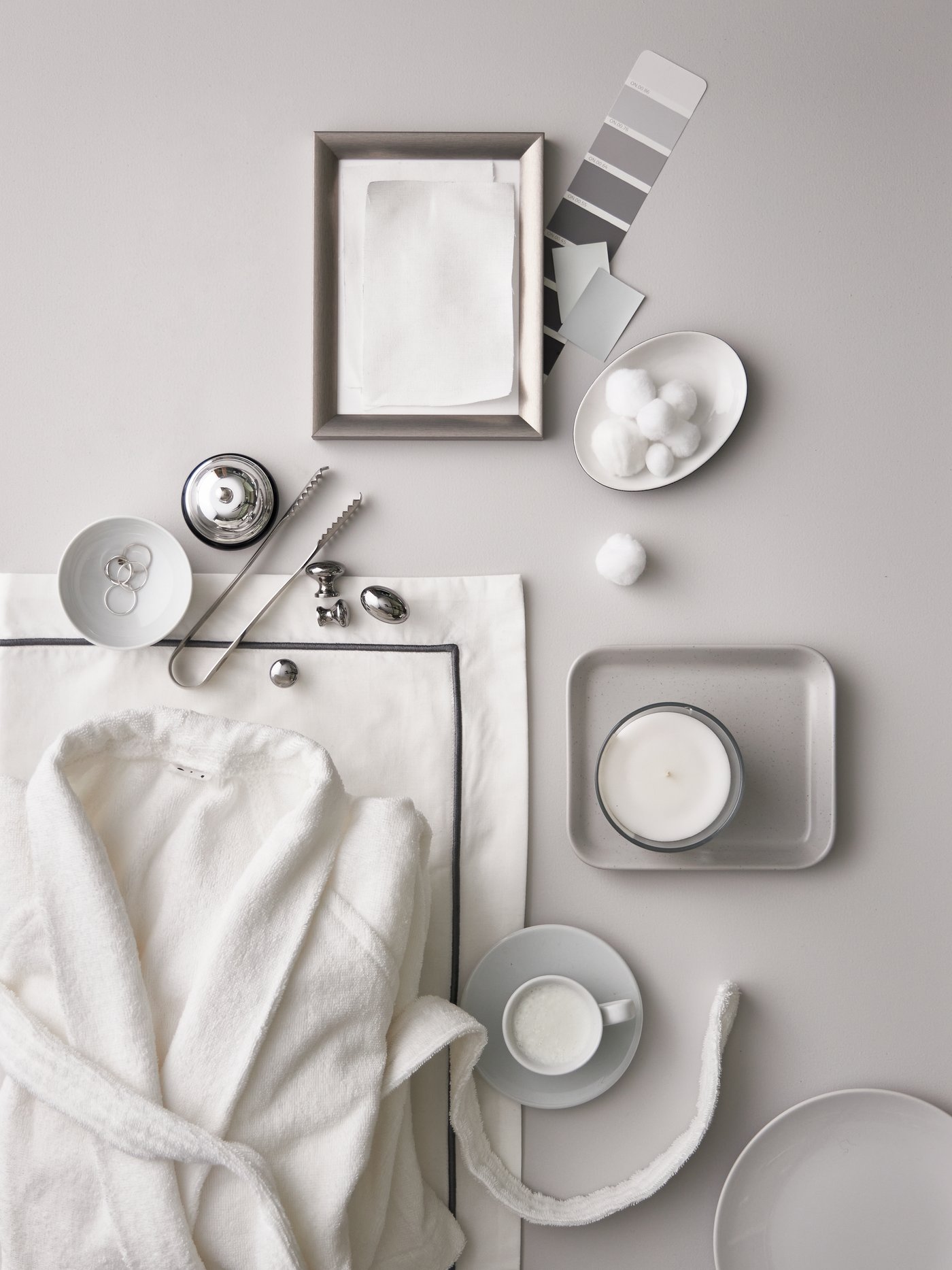 A white ROCKÅN bathrobe, white/dark grey bed linen, a small silver frame, white bowls and cups with sugar lumps and candles.