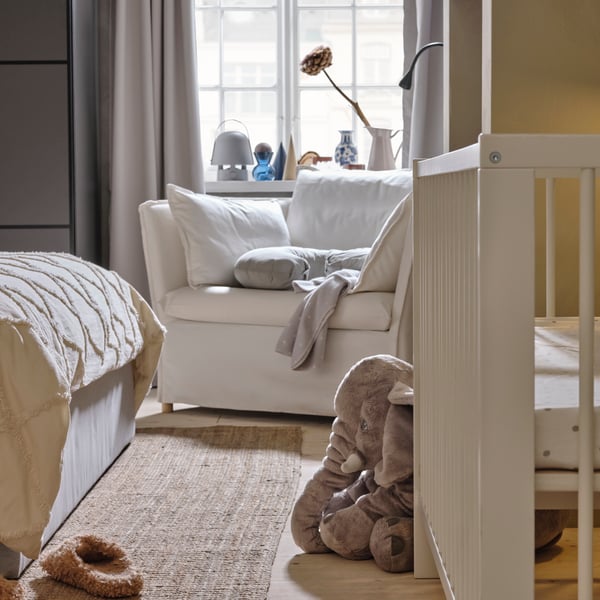 Een wit GULLIVER babybedje tegen een lichtgrijze wand bij het voeteinde van een bed. Een BACKSÄLEN 1,5-zits fauteuil staat erachter bij een raam.