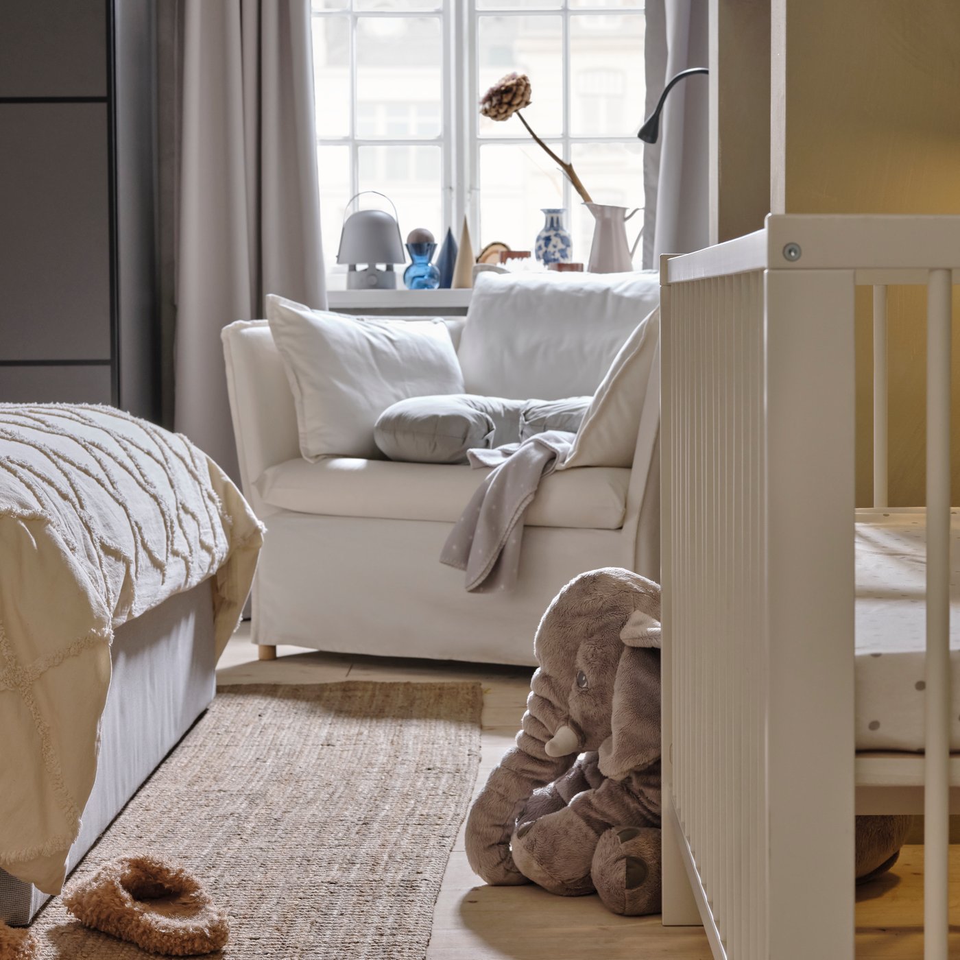 Een wit GULLIVER babybedje tegen een lichtgrijze wand bij het voeteinde van een bed. Een BACKSÄLEN 1,5-zits fauteuil staat erachter bij een raam.