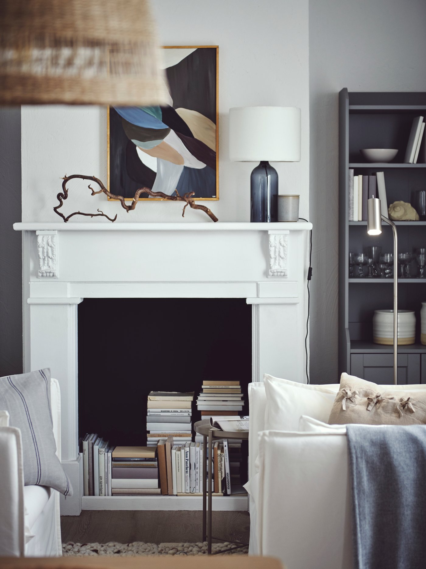 明るいリビングルームにある装飾的な暖炉の中に本が積み重ねてある。グレーのHAUGA/ハウガ ハイキャビネット、ホワイトのBACKSÄLEN/バックセーレン アームチェア。