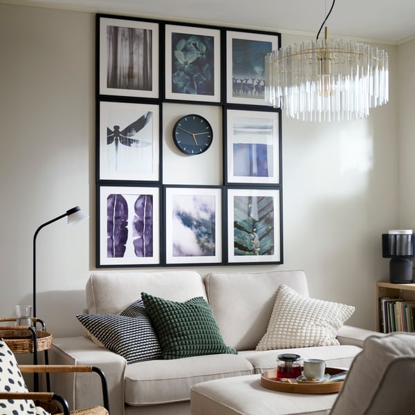 A black SKÄRIG wall clock surrounded by a symmetric arrangement of eight frames of art, hung above a KIVIK sofa.