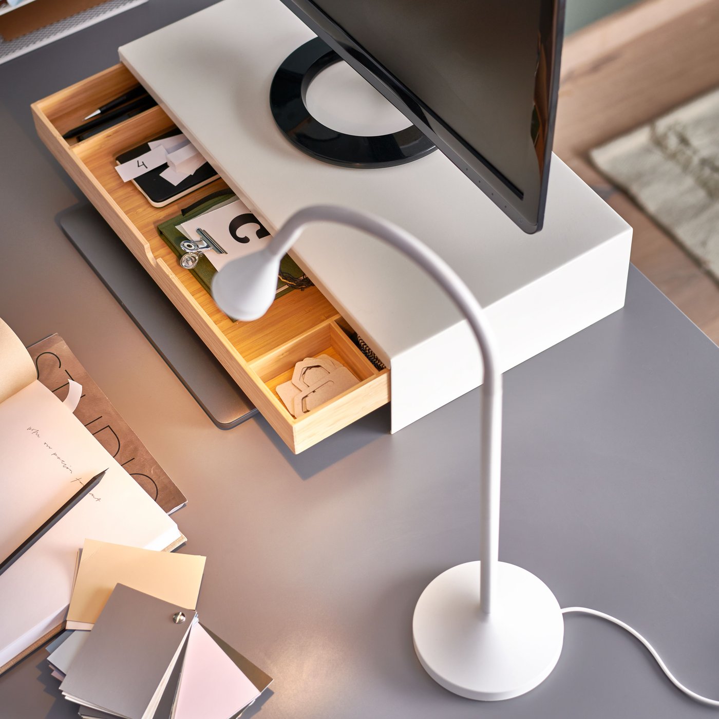 Auf einer grauen Tischplatte steht neben einer ELLOVEN Monitorerhöhung mit Schublade eine NÄVLINGE LED-Arbeitsleuchte in Weiß.