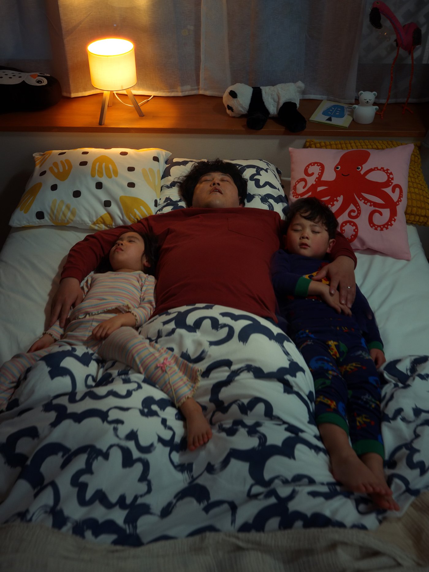 Three people lying in bed. Colorful pillows and stuffed animals in the background.